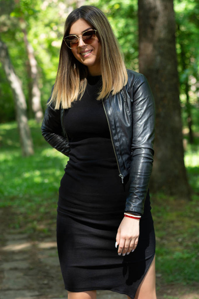glückliche Frau in schwarzem Kleid und Lederjacke und Sonnenbrille, die an einem sonnigen Frühlingstag in der Natur posiert, mit der Hand auf der Hüfte. Wald im Hintergrund. Modekonzept. Nahaufnahme, selektiver Fokus - Foto, Bild