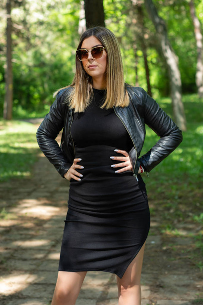 Mignon mannequin femme en robe noire et veste en cuir et lunettes de soleil posant dans la nature par une journée de printemps ensoleillée avec les mains sur les hanches. Forêt en arrière-plan. Concept de mode
 - Photo, image