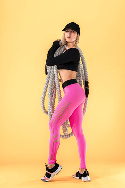 attraktive Frau in rosa und schwarzer Sportbekleidung mit schweren Seilen, die auf gelbem Hintergrund in die Kamera schauen. Stärke und Motivation. Sportliche Frau arbeitet mit schweren Seilen - Foto, Bild