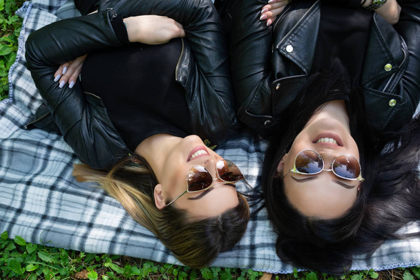 Ευτυχισμένα κορίτσια ξαπλωμένοι στην κουβέρτα και γελούν σε ένα καταπράσινο λιβάδι σε μια ανοιξιάτικη μέρα στη φύση. Τα κορίτσια φοράνε γυαλιά ηλίου και ένα μαύρο μπουφάν. Χαλάρωση και να απολαύσετε την ιδέα. Κοντινό - Φωτογραφία, εικόνα