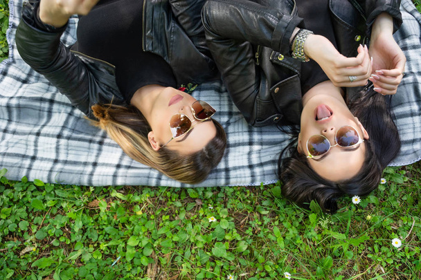 Doğada bir bahar gününde yeşil bir çayırda battaniyenin üzerinde yatan iki mutlu kız. Kızlar güneş gözlüğü ve siyah ceket giyer. Bir kız dilini dışarı soll. Yakın çekim, seçici odak - Fotoğraf, Görsel