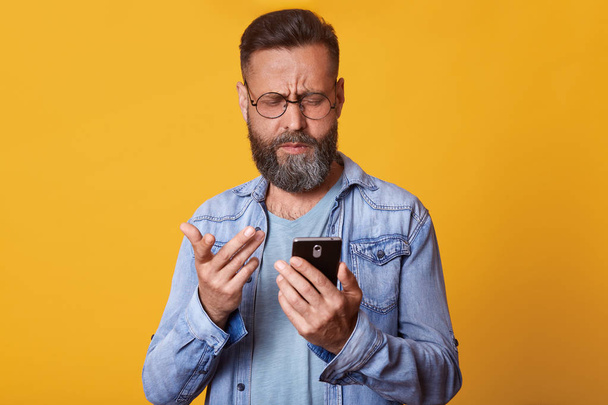 Close up portrait of single handsome Caucasian middle aged man in casual T shirt and denim jacket, texting using smartphone and has displeased facial expression, standing over yellow background. - Photo, image