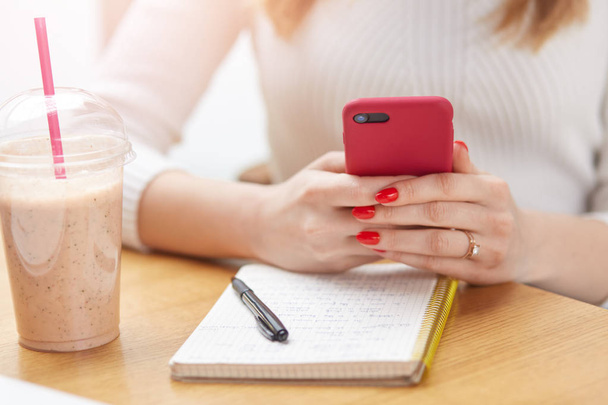 Cropped image of unrecognizable woman with red manicure, holding smart phone in both hands, making notes, having rest during lunch, drinking milkshake from plastic cup, planning her daily routine. - Foto, afbeelding