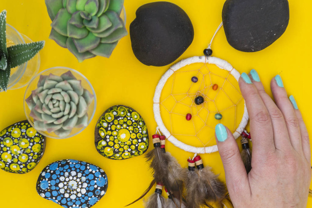An Indian dreamcatcher with feathers lies in a woman's hand among cacti and succulents on a color bright yellow background. - Foto, imagen