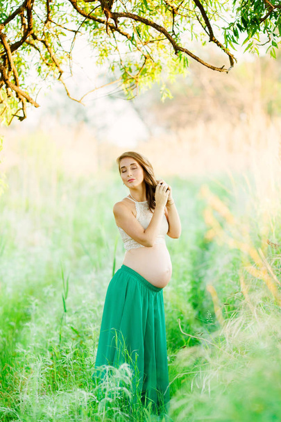 A sweet, fair-haired pregnant girl in a green skirt is standing in the park with her eyes closed and braids her braid. - Foto, Bild