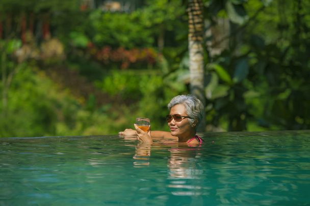 natural lifestyle portrait of attractive and happy middle aged Asian woman relaxed at tropical resort infinity simming pool with jungle background enjoying a drink relaxed - Zdjęcie, obraz