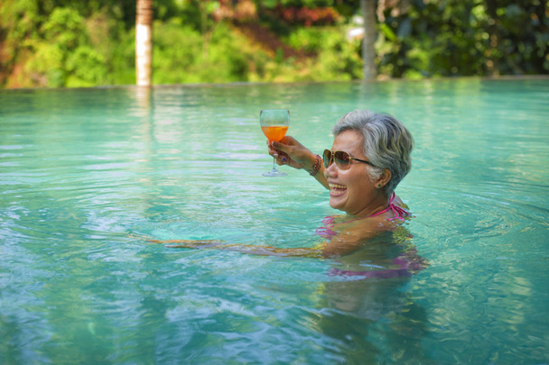 natural lifestyle portrait of attractive and happy middle aged Asian woman relaxed at tropical resort infinity simming pool with jungle background enjoying a drink relaxed - Photo, Image