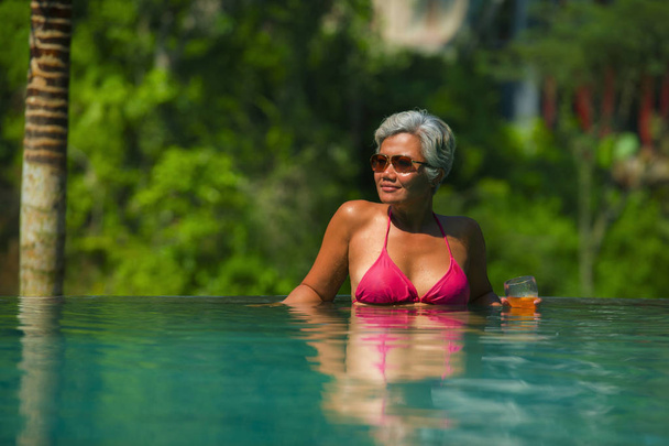 natural lifestyle portrait of attractive and happy middle aged Asian woman relaxed at tropical resort infinity simming pool with jungle background enjoying a drink relaxed - Photo, Image