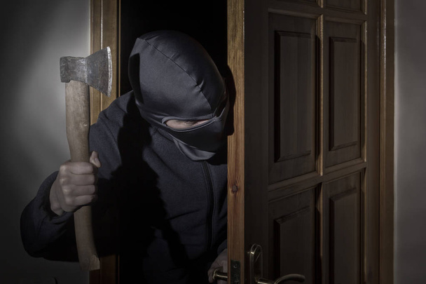 Ladrão com um machado na mão e em um chapéu com uma fenda para os olhos foge para dentro de casa através da porta à noite ao luar
 - Foto, Imagem