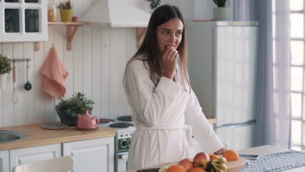Cute girl in white bathrobe eats slice of orange in kitchen, slow motion - Video
