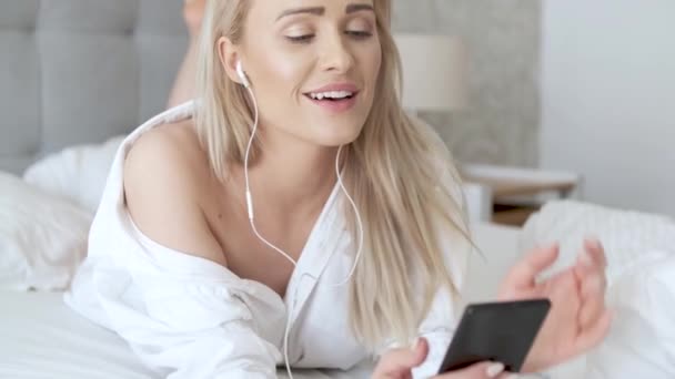 Beautiful, smiling blond woman lying in white bed and using a smartphone - Imágenes, Vídeo