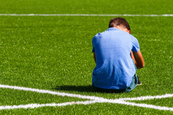 Smutný zklamaný chlapce, který seděl na trávě stadionu - Fotografie, Obrázek