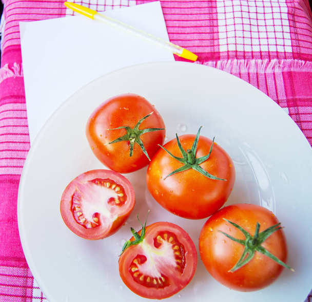 Fresh ripe red tomatoes and sliced tomatoes with water drops and a green peduncle on a white plate, next to a sheet of Notepad for controlling diet - Photo, Image