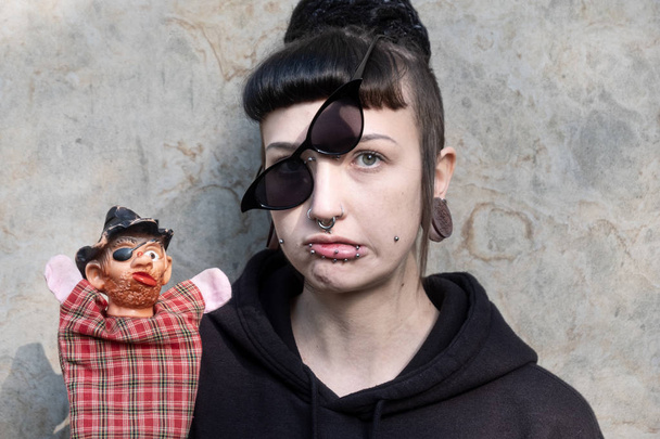 Beautiful young individual, eccentric woman, with interesting hairstyle and piercings and obliquely put on glasses questioningly shows a hand puppet. - Foto, afbeelding