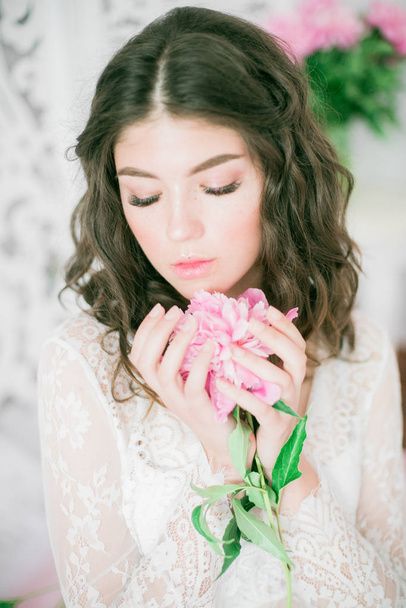 Beautiful young girl in a white lace dress with peony flowers in a bright studio - Photo, Image