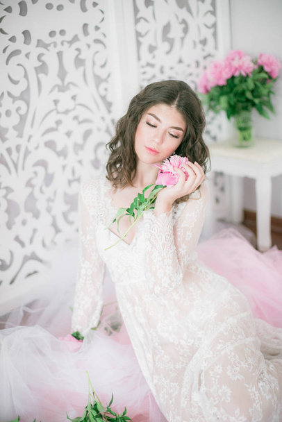 Beautiful young girl in a white lace dress with peony flowers in a bright studio - Fotó, kép