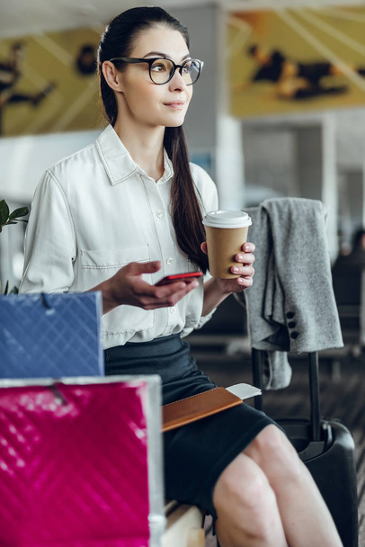 Serene elegant woman with phone is drinking coffee in airport - Photo, Image