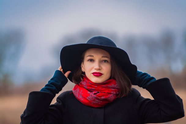 Tyttö mustassa takissa, lapiohattu päässä ja punainen huulipuna huulilla, kävelee puistossa. Tyyli ja moderni muoti
. - Valokuva, kuva