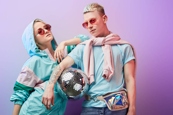 Young fashionable couple of dancers posing with disco ball on violet background - Foto, imagen