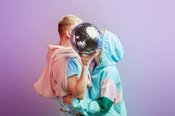 Young fashionable couple of dancers posing with disco ball on violet background - Фото, зображення