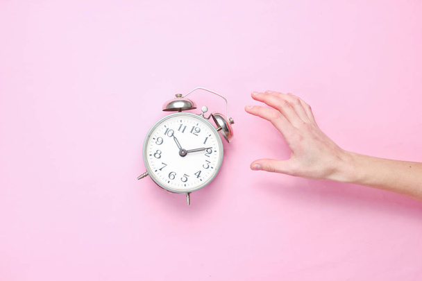 Vrouwelijke hand neemt wekker op roze achtergrond. Bovenaanzicht. Tijd om wakker te worden. Minimalis - Foto, afbeelding