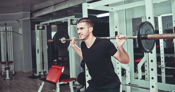 Training at gym. Young man crouches with barbell on the shoulders side view - Photo, Image