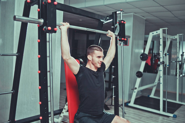 Young sports guy trains the muscles of the shoulders (deltas) with a bench press while sitting in a gym machine. Training proces - Fotoğraf, Görsel
