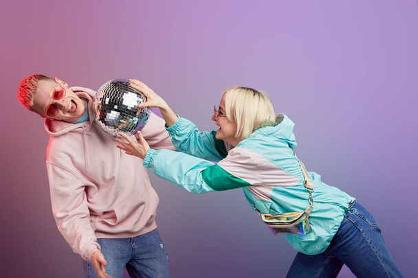 Young fashionable couple of dancers posing with disco ball on violet background - 写真・画像