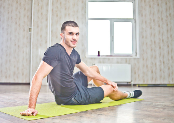 Uomo sportivo sorridente alla macchina fotografica mentre si siede sul tappeto yoga e si riposa dopo l'esercizio
. - Foto, immagini