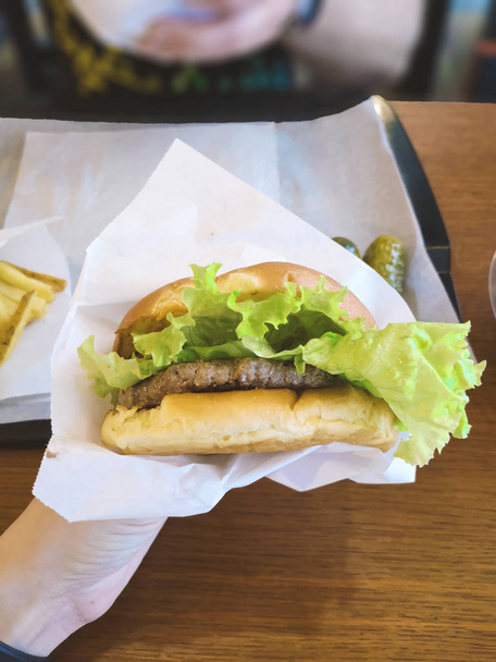 hamburger holding female hand in paper craft packaging in a fast food cafe with lettuce leaf, medium roast meat patty, tomato next to lettuce in a plastic container, unhealthy no diet food - Photo, Image