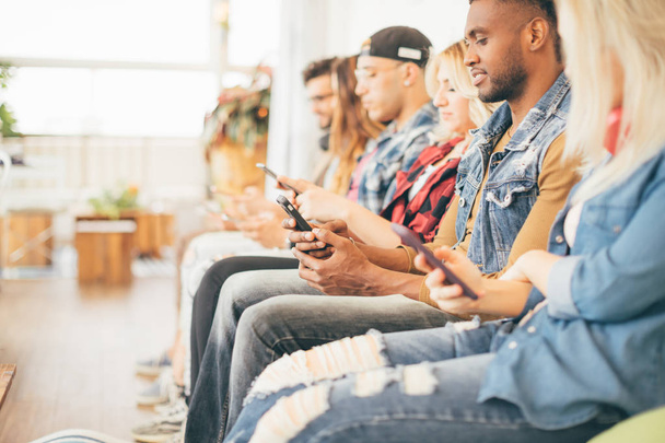 Picture of multiracial group of young people sitting with smartphones in their hands, looking into screens and smiling - technology and social media addiction concept - Foto, Imagem