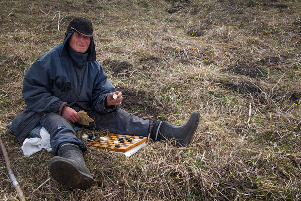 Un anciano con ropa desordenada y una gorra se sienta en una colina jugando ajedrez en la primavera contra el telón de fondo de la naturaleza marchita
 - Foto, Imagen