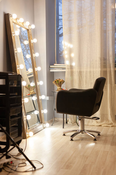 Workplace stylist, hairdresser, barber, makeup artist. Beauty saloon. Black chair for client opposite large floor mirror with bulbs, organizer for tools. Evening time, cozy atmosphere. - Fotografie, Obrázek