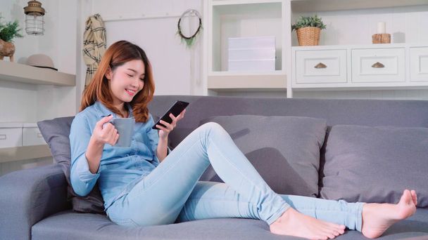 Portrait of beautiful attractive smiling Asian woman using smartphone holding a warm cup of coffee or tea while lying on the sofa when relax in living room at home. Lifestyle women at home concept. - 写真・画像