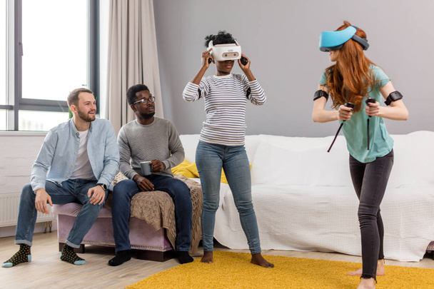 Two multiracial young women dancing wearing virtual reality glasses while their boyfriends sitting on couch and smiling - Photo, Image