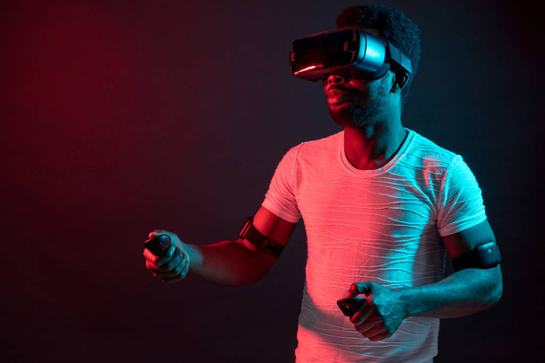Young man wearing VR headset watching video, isolated on dark red dual light. - Φωτογραφία, εικόνα