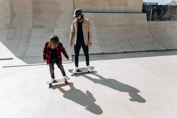 Young father and his son ride skateboards in a skate park with slides outside at the sunny day - Zdjęcie, obraz
