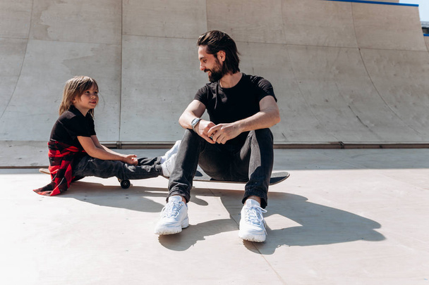 Jeune père et son fils habillés dans les vêtements décontractés sont assis sur les planches à roulettes dans un skate park au soleil jour
 - Photo, image