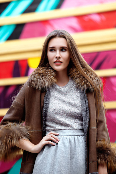 Fashionable long-haired girl dressed in a gray dress and a long brown sheepskin coat poses in the street - Foto, Bild