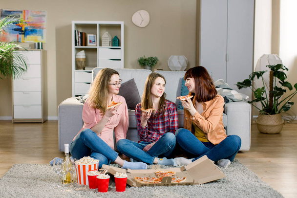 friendship, holidays, fast food and celebration concept - happy young women friends with drinks and popcorn eating pizza at home, sitting on the floor in cozy room - Photo, image