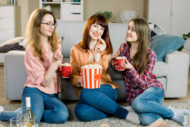 Women friendship, home party. Three beautiful funny young girl friends with popcorn in hands sitting on the floor in cozy room at home and looking on camera - Foto, Bild