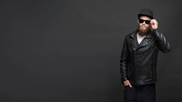 Man wearing leather biker jacket or asymmetric zip jacket with black cap, jeans and sunglasses. Handsome hipster man over gray background with copy space - Valokuva, kuva