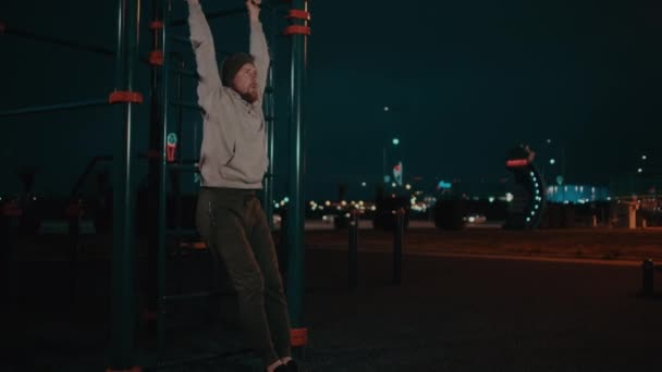 Bearded guy is hanging on training area in park in night and lifting legs - Filmati, video