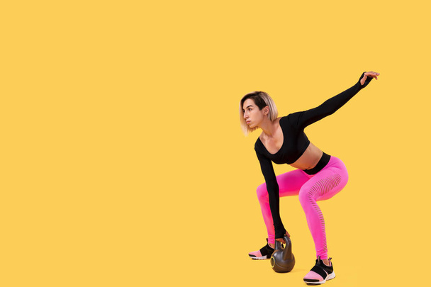 Fitness woman in fashionable pink and black sportswear work out with kettlebell on yellow background. - Zdjęcie, obraz