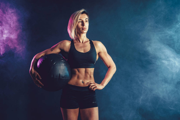 Sporty blonde woman in fashionable sportswear posing with medicine ball. Photo of muscular woman on dark background with smoke. - Photo, Image