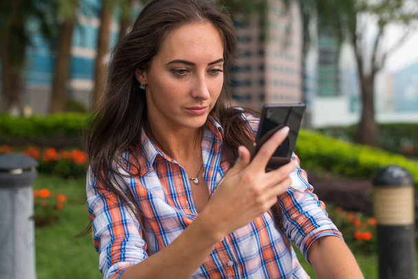 junge Frau, die draußen an einem Tisch sitzt und ihr Handy benutzt. Frau liest SMS auf Smartphone im Park. - Foto, Bild