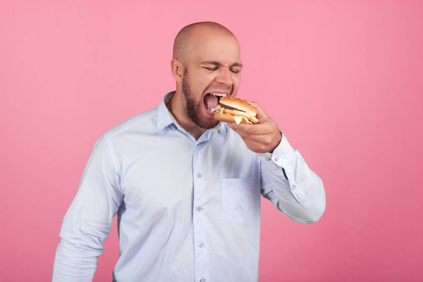 Spokojený muž se svěží bradkou a plešatou hlavou. otevírá velmi širokou pusu, aby si co nejdříve snědl hamburger. Postavte se před růžové pozadí - Fotografie, Obrázek