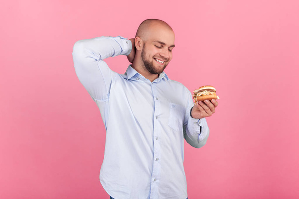 A satisfied man with a lush beard and a bald head. looks at his burger with a thirst for him to eat it as soon as possible. stand in front of the pink background - Foto, immagini