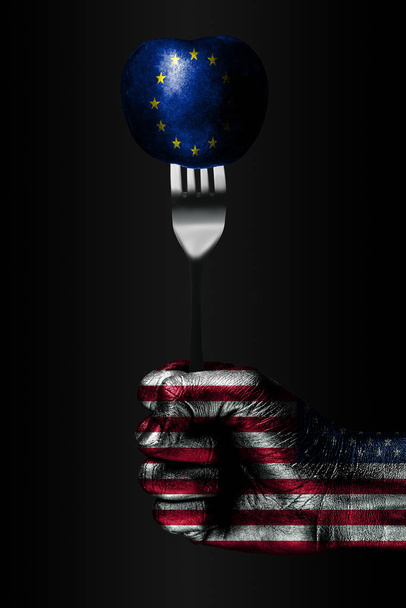 A hand with a drawn USA flag holds a fork, on which is a ball with a drawn EU flag, a sign of influence, pressure, grip and anecxia. - Photo, Image