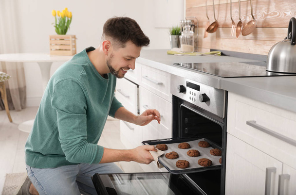 Handsome man taking out tray of baked cookies from oven in kitchen - Фото, изображение
